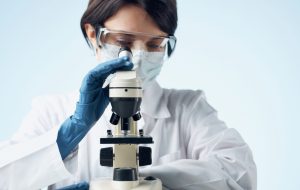 beautiful woman HGH Doctor in glasses and in medical mask blue gloves on hands and microscope on the table  300x190